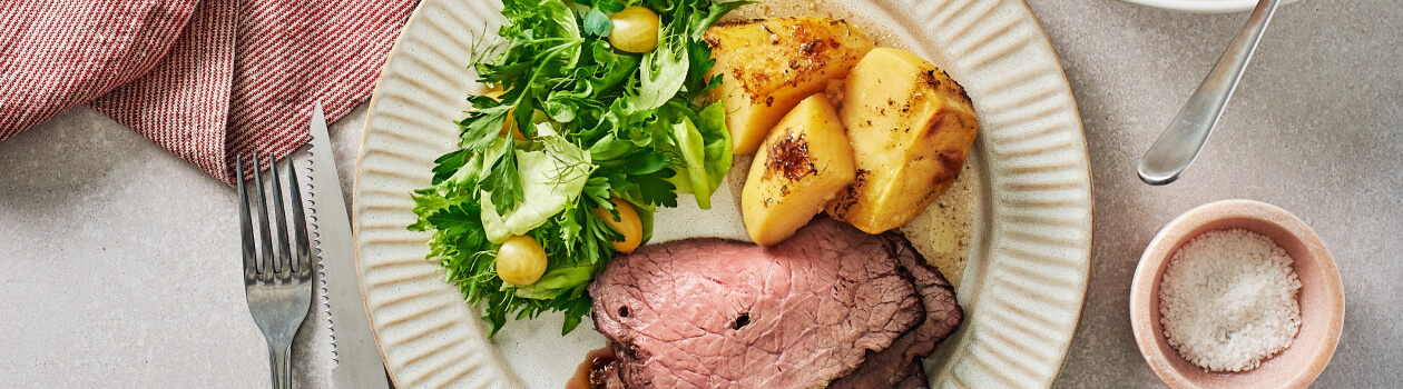 Sliced roast on a white plate with a side of green salad and potatoes beside the full roast sliced on a cutting board.
